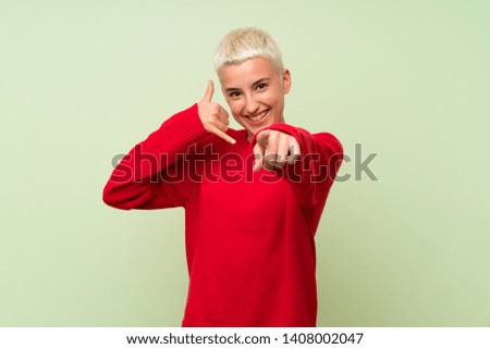Teenager girl with white short hair over green wall making phone gesture and pointing front