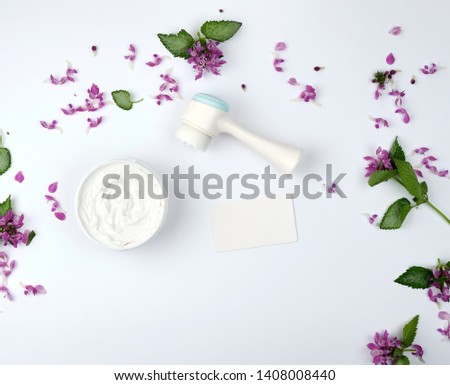 white moisturizing thick cream in a plastic jar, massager and empty paper white business card, white background with flowers and green leaves, top view
