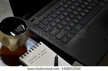 Picture of a cup of coffee in focus near a laptop and notepad.