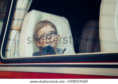 A little boy with round glasses looks through the long distance bus window with raindrops on it. Child leaving for vacation or holidays. (vintage processing, noises, film photo)