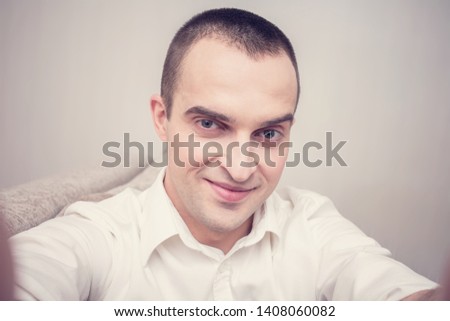 Attractive young guy in white shirt smirking for selfie, sitting at home on the couch, front view, toned