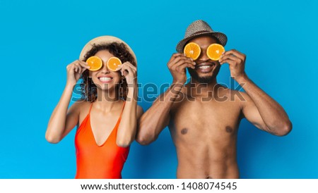 Cheerful young black couple holding two halves of orange near eyes, blue studio background. Healthy summer concept, panorama