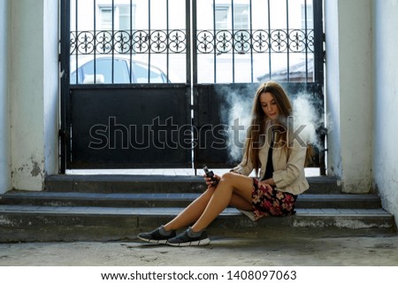 Vape teenager. Young cute girl in casual clothes smokes an electronic cigarette near the wall in prison cell. Bad habit that is harmful to health. Vaping activity.