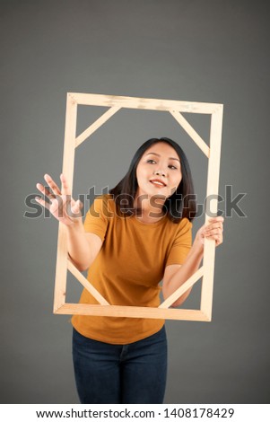 Young Asian woman holding wooden picture frame and stretching out her hand to take something