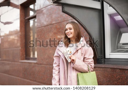 Young woman with money and shopping bag near cash machine outdoors