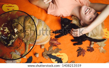 Beautiful young surprised woman in witches hat and costume holding pumpkin. Black cat with pretty girl. Beautiful young woman in black witch costume with hat over orange background
