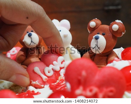  hands hold a red heart with a teddy bear on a birthday cake on a blurred background.