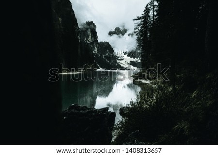 view on a mountain lake up high in the italian dolomites with the lake in the centre surrounded by dark green trees and snow covered mountains in the background reflecting in the lake in a moody edit