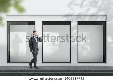 Smiling businessman with phone walking near white building with three vertical mock up posters in windows. Concept of advertising and marketing.