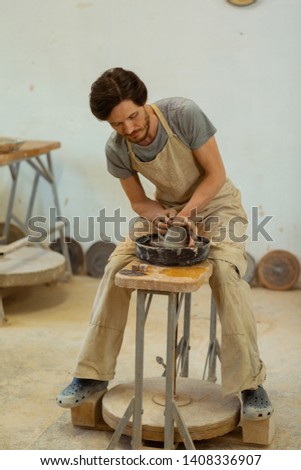 Producing new creation. Dark-haired man with beard manufacturing new clay pots while using professional wheel