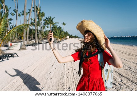 woman in a hat with a backpack in a red dress holding a phone near the beach ocean palm trees                              