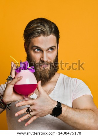 Attractive, stunning and cheerful bearded young man smiles and holds a multicolored cocktail with a straw. looking away. free space for your advertising. On a yellow background.