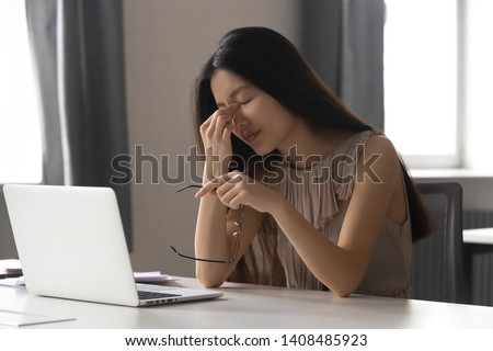 Overworked stressed asian business woman employee holding glasses feeling pain in dry eye strain or headache suffer from bad blurry weak vision tired of computer work syndrome, sight problem concept Royalty-Free Stock Photo #1408485923