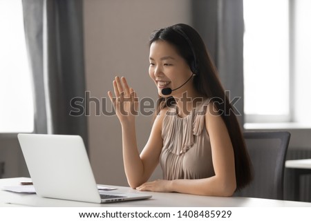 Smiling asian businesswoman wearing wireless headset waving hand doing video chat looking at laptop consulting customer sitting at workplace, female call center service support agent talking  Royalty-Free Stock Photo #1408485929