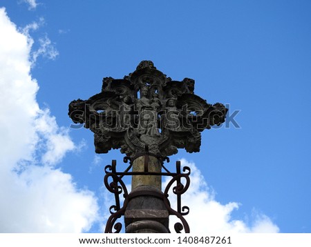 Decorative heavy dark stone cross designed as a flower with floral patterns and the Virgin Mary and baby Jesus in her arms at its heart flanked by two saints against blue sky background. France
