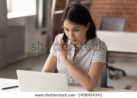 Serious asian businesswoman using laptop looking at computer talking on phone consult client sit at desk, focused female sales manager make mobile business call discuss work with customer in office Royalty-Free Stock Photo #1408494341