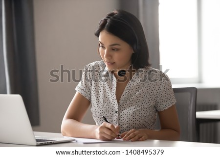 Serious asian businesswoman wear headset watching video webinar make conference
 online call write notes, focused chinese woman study online looking at laptop working in customer service support 