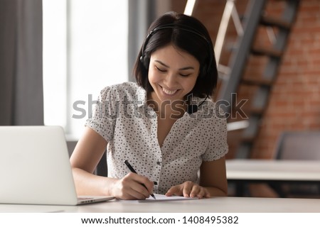 Happy asian businesswoman wearing headset make notes talk by conference call doing video chat on laptop in office, smiling chinese support service agent write information consult customer Royalty-Free Stock Photo #1408495382