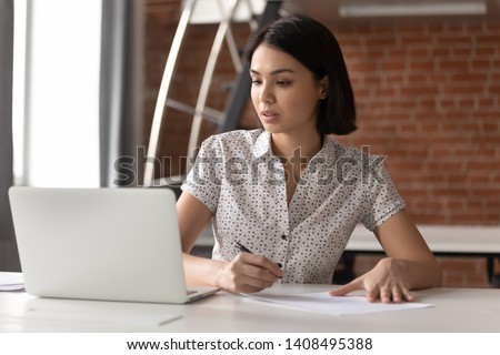 Focused asian business woman working studying online in office looking at laptop making notes, serious japanese employee or student watching webinar writing information in notebook sitting at desk Royalty-Free Stock Photo #1408495388