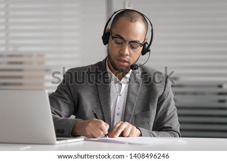 Serious african male customer support service agent wear wireless headset talk write notes make video conference call, focused mixed race businessman operator representative  online chat