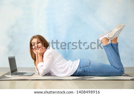 Beautiful young woman with laptop lying on floor against color wall