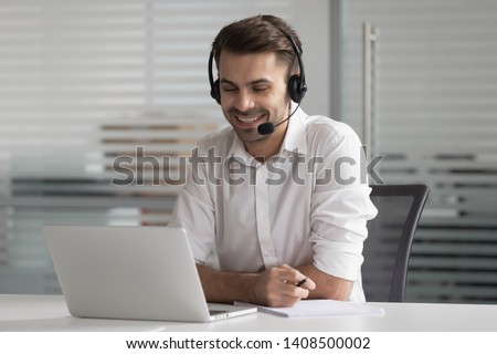 Smiling business man wearing wireless headset make conference video call looking at laptop screen, happy salesman customer service support agent helpline representative talk by online chat in office Royalty-Free Stock Photo #1408500002