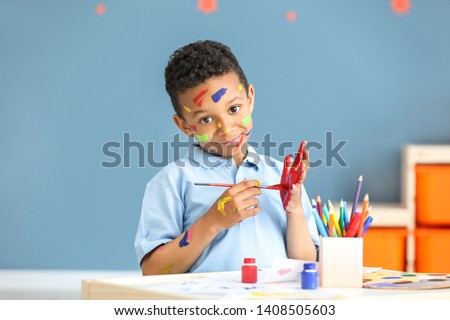 Cute African-American boy with hands and face in paint at home