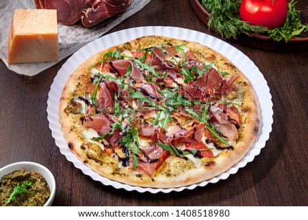 Top view on pizza with tomato, parmesan and prosciutto with white sauce with ingridients on brown wooden table. Picture for recipe or menu. Copy space for design. Served cooked pizza. Italian cuisine
