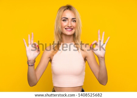 Young blonde woman over isolated yellow wall showing an ok sign with fingers