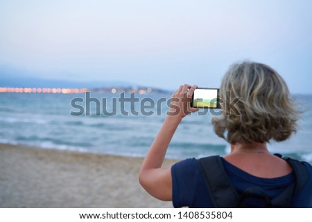 Woman taking a photo of a sea on smart phone on sunset at the beach, back view. Focus on phone