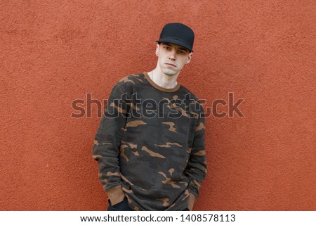 Cute handsome young man in a stylish black cap in a trendy green military shirt in jeans posing in the city near a vintage building pantone color 2019. American cool guy model. Trendy menswear.