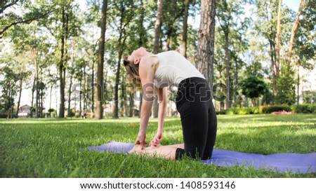 Photo of smiling middle aged woman in fitness clothes doing stretching and yoga exercises. WOman meditating and doings sports on fitness mat on grass at park