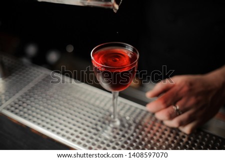Bartender pouring a delicious red cocktail from the measuring glass cup on the bar counter in the dark blurred background