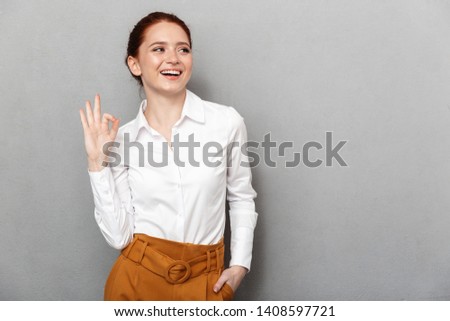Portrait of european redhead businesswoman 20s in formal wear smiling at camera and showing ok sign in office isolated over gray background
