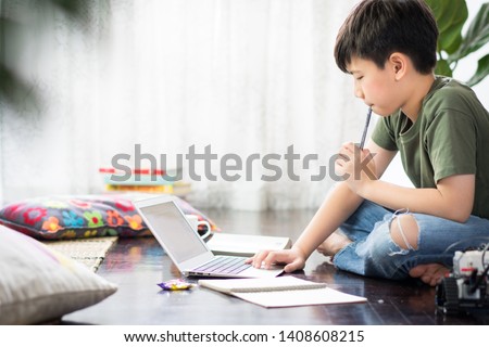 Smart looking Asian preteen boy sit crossed legs, holding pen against his lips, thinking and look at computer notebook at home due to Covid-19 pandemic and social distancing, New normal, Remote class. Royalty-Free Stock Photo #1408608215