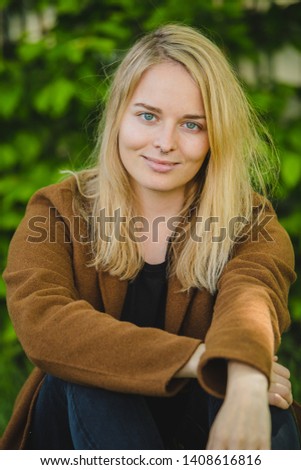 Young blonde hipster girl seductevly looking and smiling towards the camera with her arms crossed on her leg. Green bush in the background.
