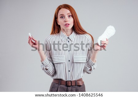 Closeup photo of young redhead businesswoman in striped shirt holding hygiene pad choosing tampon studio white background