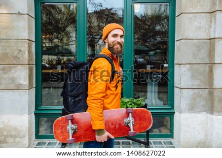 Portrait of stylish positive beard hipster man traveler wearing a orange hat and jacket walking along the street with a skateboard or longboard in his hand. Lifestyle outdoor concept.