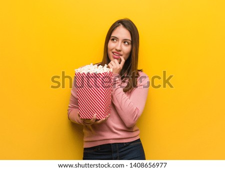 Young cute woman holding a popcorn bucket relaxed thinking about something looking at a copy space