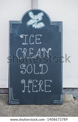 Close up of a black "Ice cream sold here" chalkboard sign leaning against white wall.
