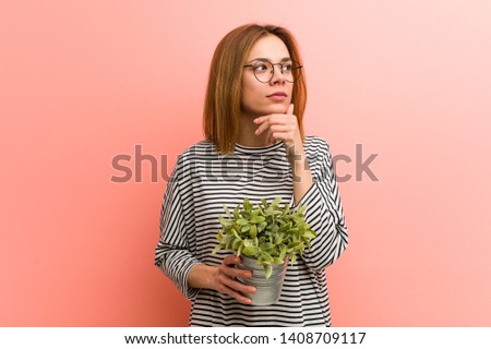 Young woman holding a plant looking sideways with doubtful and skeptical expression.