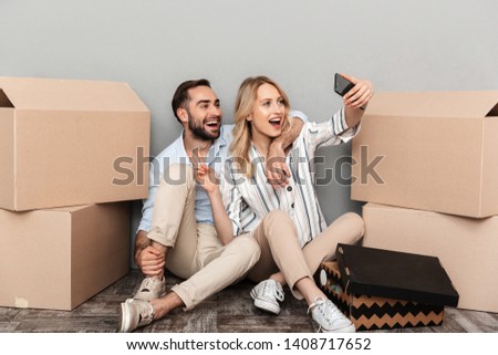 Photo of positive couple seating near cardboard boxes and taking selfie photo on cellphone isolated over gray wall