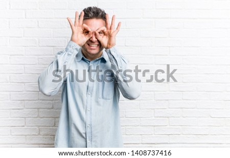 Young handsome man against a bricks wall showing okay sign over eyes