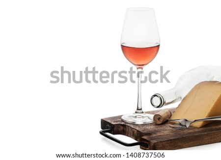 An overhead view of a glass of rose wine, an empty bottle, a cork, some hard cheese and a dessert fork located on an old cutting board with a metal handle in a white background