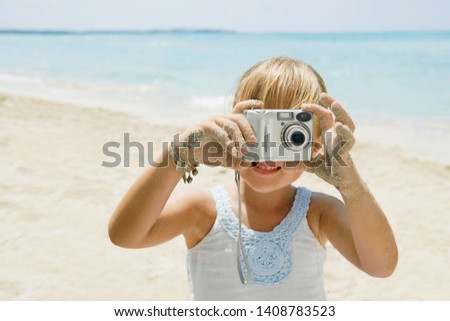 Beautiful child girl holding digital photo camera on beach summer holiday, taking pictures sunny outdoors. Family vacation memories, taking photos using technology, leisure recreation lifestyle.