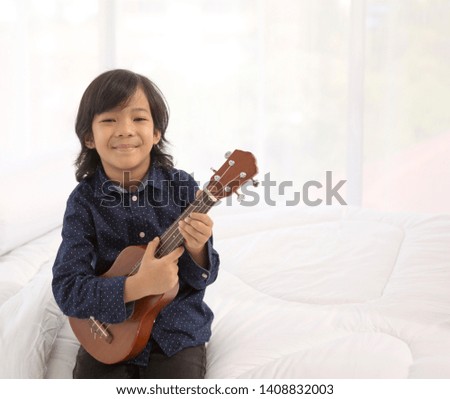 A cute little smiling asian boy is playing his ukulele on the bed