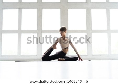 Young attractive smiling woman practicing yoga, sitting in One Legged King Pigeon exercise, Eka Pada Rajakapotasana pose, working out, wearing sportswear, brown pants, bra, indoor full length, home