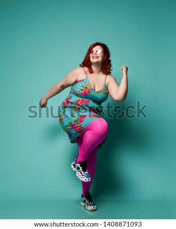 Plus-size lady overweight woman in fashion sunglasses and colorful clothes shows Yes sign with her arm, dances funny, do excercises and have fun on mint background