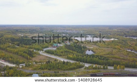 Aerial view of bright green fields, small pond, houses and a road with moving cars. Clip. Natural landscape with meadow, pine trees on cloudy sky background.