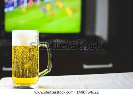 Mug of beer in front of  television, with football-soccer game in background. Fresh cold beer in living room.
